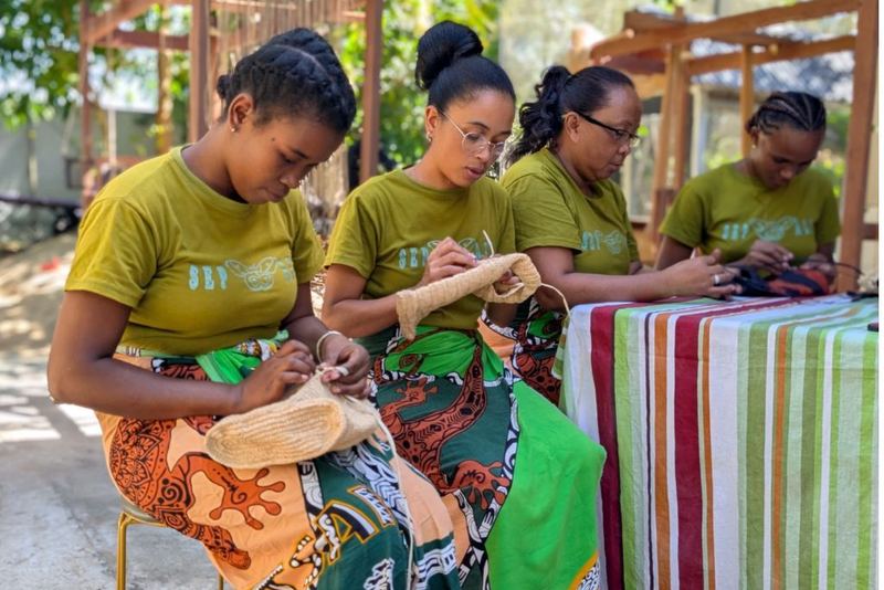 Four artisans in green shirts weaving raffia beside a striped textile.