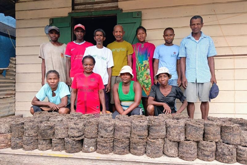 A village group standing behind rows of fuel bricks pressed from invasive plants.